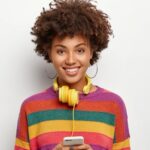 delighted-african-american-teenage-girl-holds-mobile-phone-connected-headphones-min